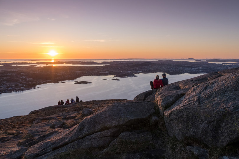 Et par sitter i solnedgangen på en fjellside og ser utover utsikten mot himmel, hav og begyggelse.