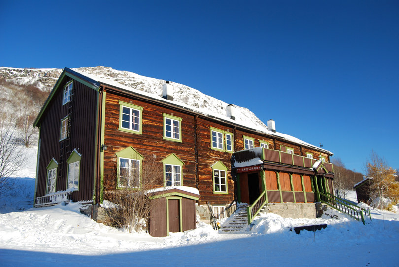 Gammelt brunt gårdshus ligger ved foten av et fjell. Sola stråler, og det er masse snø og vinter.