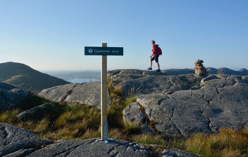 En dame går over et fjell med utsikt. Et turskilt i front hvor det står Fjogstadnuten.