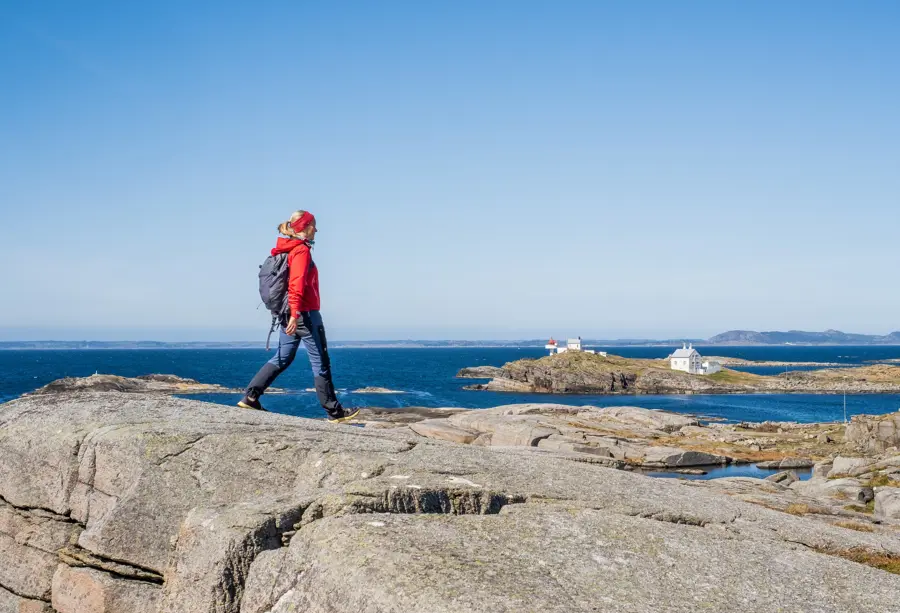 En kvinne i rødt går tur på et berg. Et fyrtårn i vises i bakgrunnen og blått hav
