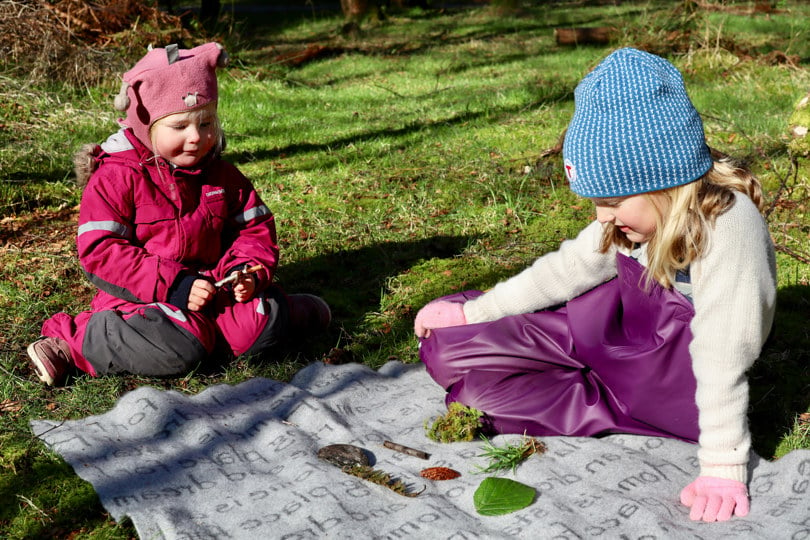 To jenter sitter på et pledd og ser på gjenstander fra naturen og leker Kims lek.