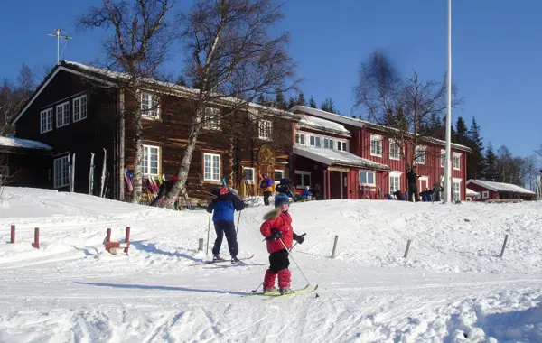 En brun og rød langstrakt hytte på vinteren, bakken rundt dekket av snø. Foran er det to barn på ski.
