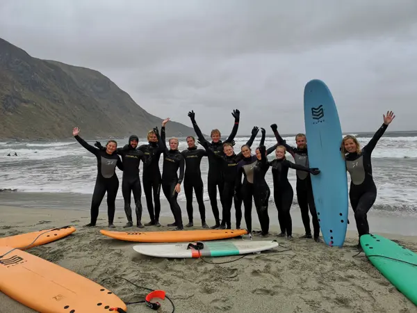 Ungdommer i våtdrakt står på strand og jubler med surfebrett liggende rundt omkring.