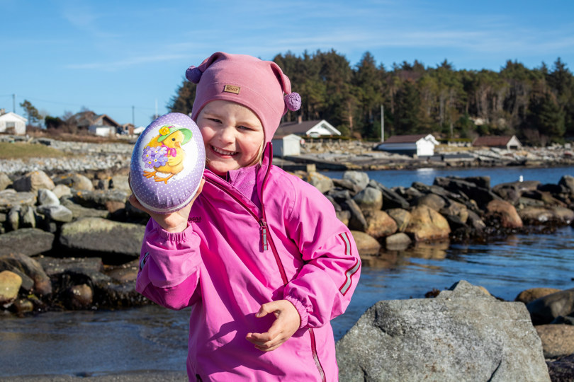 Et barn i rosa dress holder fram et påskeegg og smiler godt.