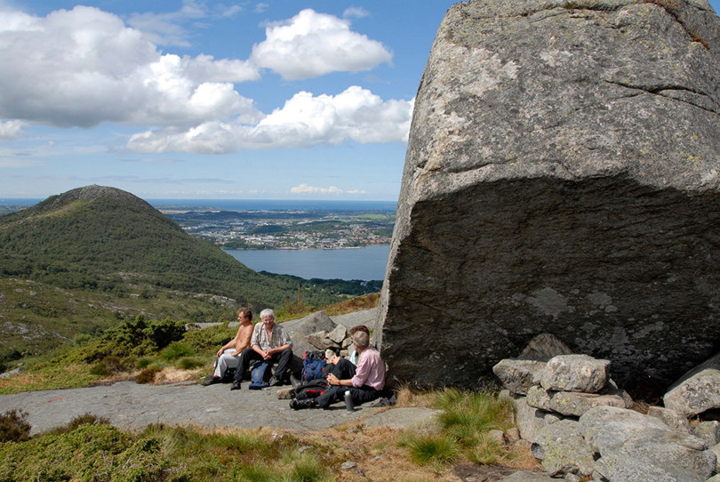 En gruppe mennesker raster ved en gigantisk stein som er kalt Resasteinen. Utsikt mot sjøen i bakgrunnen.