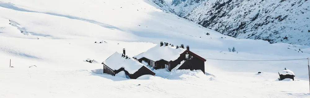 En fjellhytte dekket av snø ligger i et snødekt landskap med fjell i bakgrunnen.