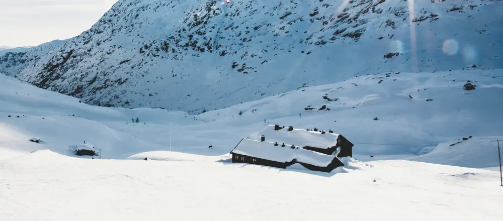 En fjellhytte dekket av snø ligger i et snødekt landskap med fjell i bakgrunnen.