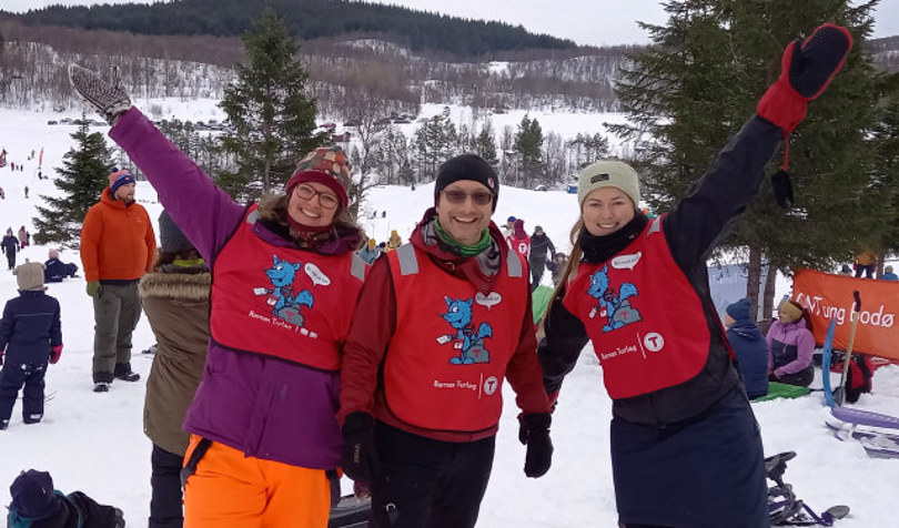 Tre voksne står smilende sammen, to av dem med armen i været. Det er snø, trær og andre folk i bakgrunnen. De har på seg fargerike klær og røde vester.