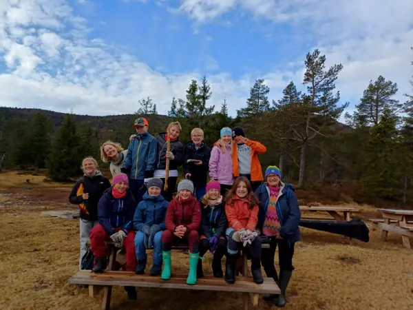 Barn og voksne sitter på en benk og smiler til kamera, bak er det skog.
