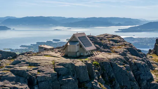 Trekantet hytte på toppen av flatt fjell med hav og øyer i bakgrunnen.