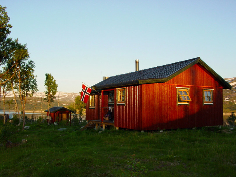 Ei rødmalt hytte med svart tak. Gule vinduskarmer. Et norsk flagg henger ved døra. Et uthus og et vann i bakgrunnen. Noen løvtrær ved siden og gress foran.