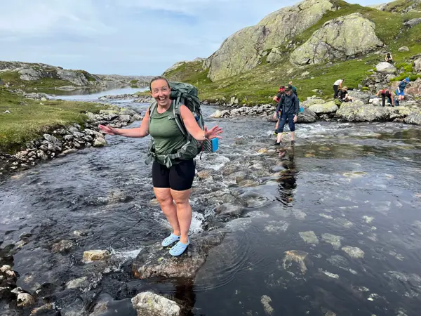 En dame krysser en elv med crocs på. Hun smiler til kamera og holder armende ut.