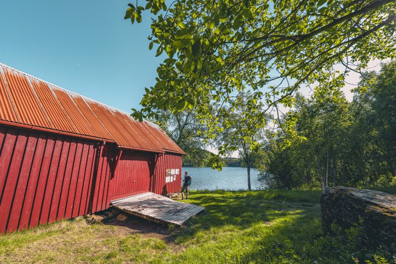 Et rødt naust foran et vann. Det er skog på andre siden av vannet. En person står ved naustet. Det er sommer og sol.
