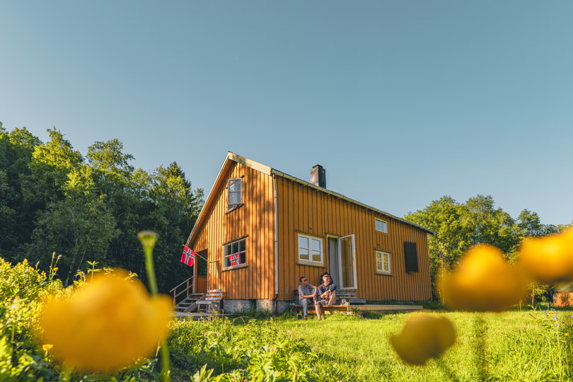 Ei gul hytte med gule blomster og gress foran. Skog i bakgrunnen. Det er sol. To personer sitter på trappen foran hytta. Hytta har et flagg ved inngangen.