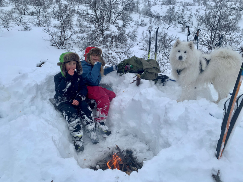 To barn og en hund er samlet rundt et bål i snøen. De spiser noe.