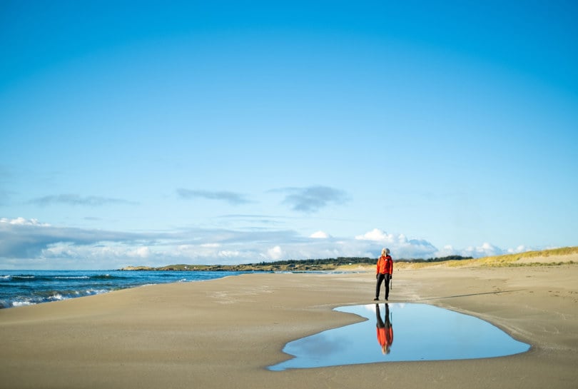 En kvinne i rød jakke står på en sandstrand med blått hav.