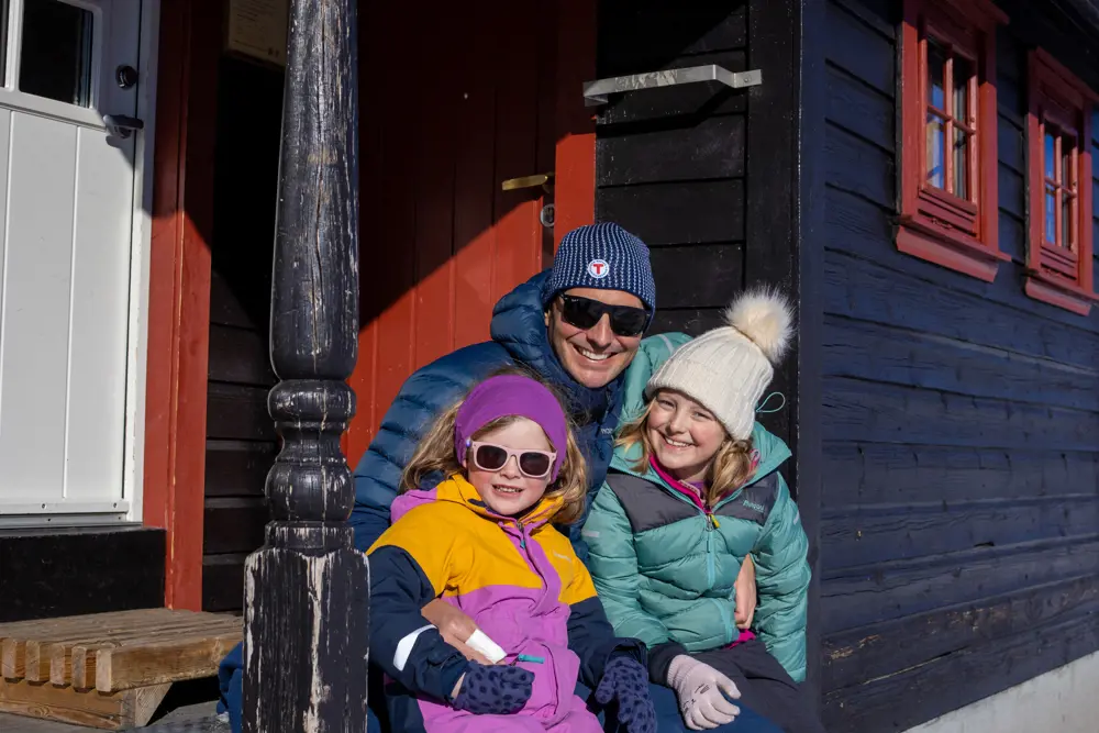 En mann og to barn sitter utenfor en svart hytte med røde karmer.