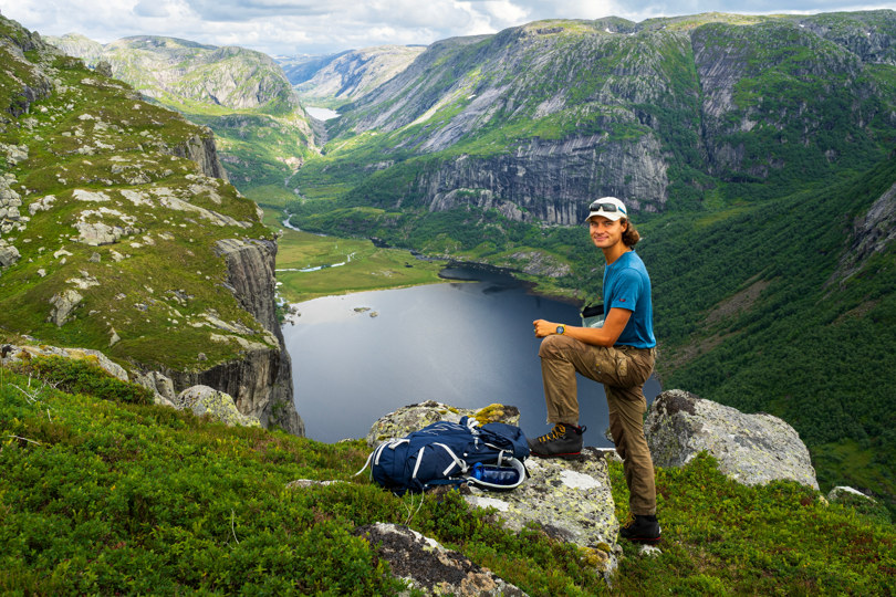 En ung mann står på kanten av et fjell. En irrgrønn dal med et vann vises i bakgrunnen.