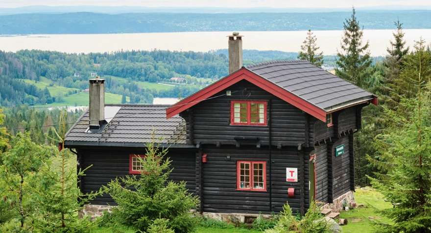 Svartmalt hytte med røde karmer med utsikt mot en fjord