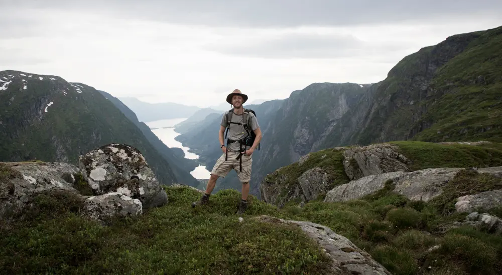 Stilig mann i knalltøff hatt på fjell med fjord bak seg.