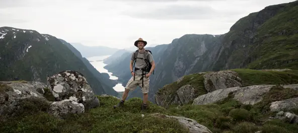 Stilig mann i knalltøff hatt på fjell med fjord bak seg.