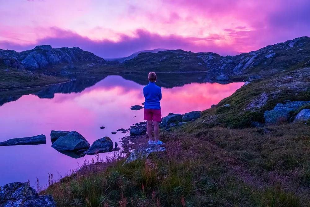 Menneske som står på et berg ved et vann mens solnedgangen lager himmelen rosa og lilla.