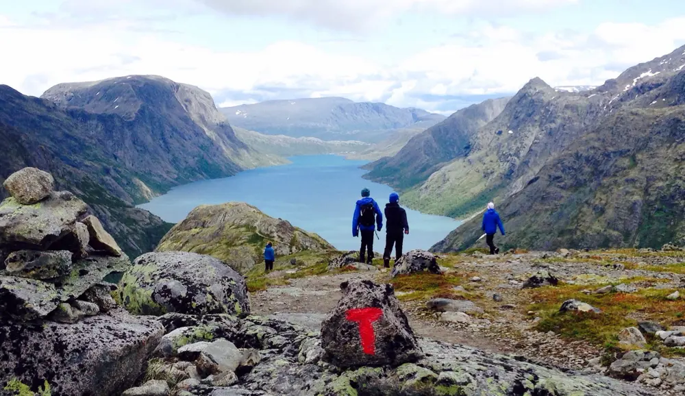 Personer og T-merket stein på fjelltopp, med utsikt mot vann og fjell i bakgrunnen