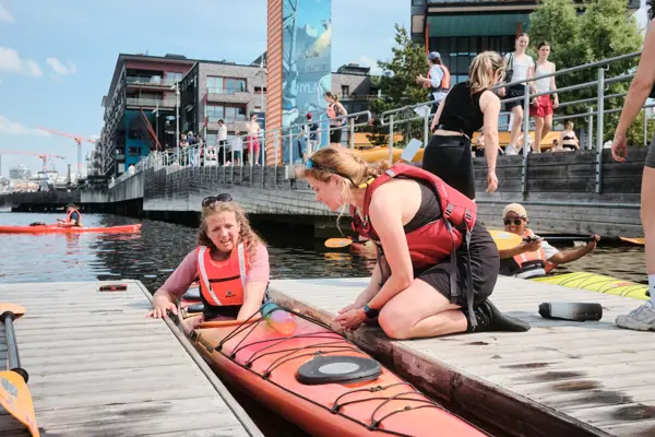 En person som sitter på en brygge og hjelper en annen person i en kajakk