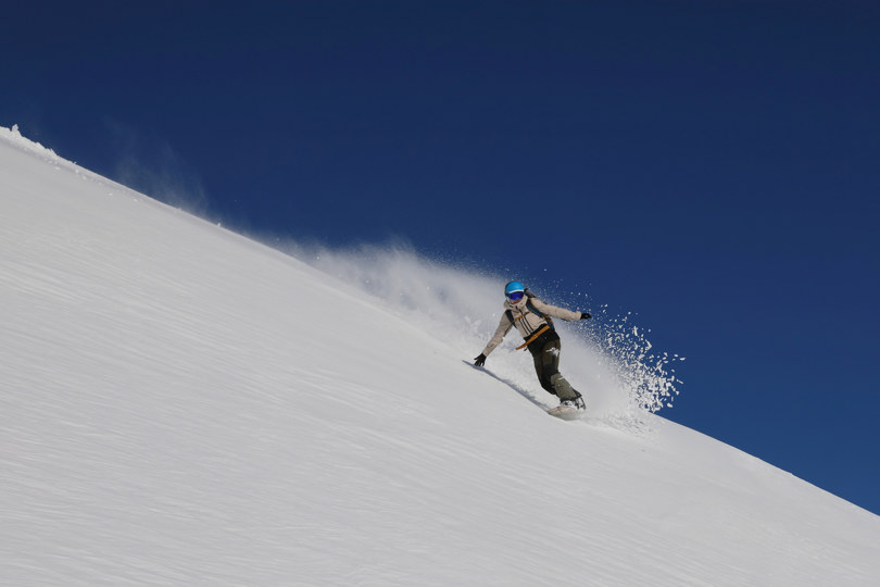 Dame på snøbrett på vei nedover en fjellvegg med snø som henger som et slør etter bak