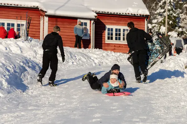 To jenter som smiler på akebrett ned bakke med rød huyttevegg bak dem.