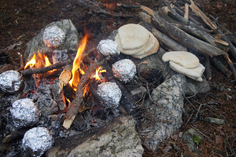 Et pinnebål mellom noen steiner. Det ligger mange runde pakker aluminiumsfolie på glørne. Ved siden av bålet ligger noen kvister og noen pitabrød.