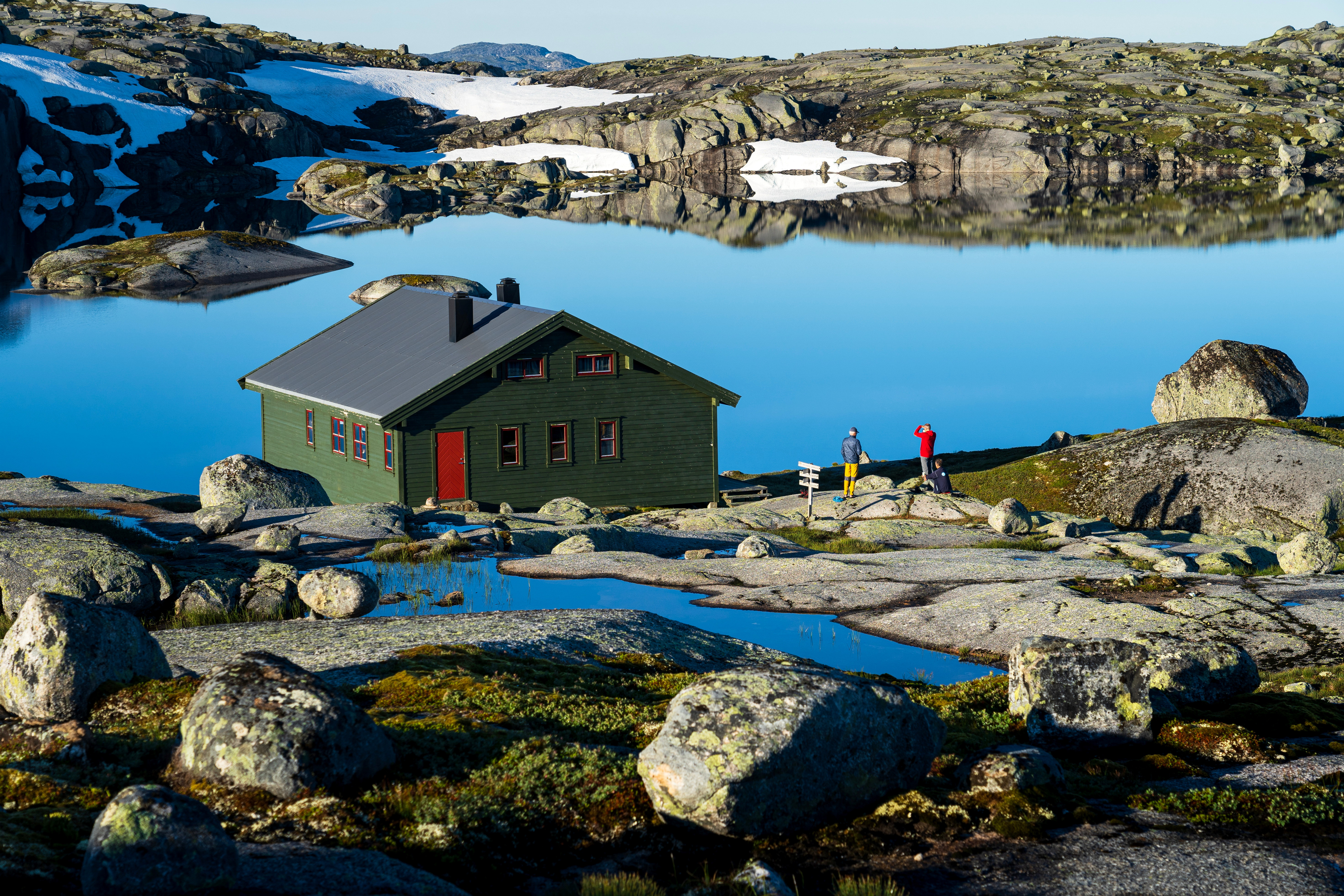 En grønn hytte som ligger ved et vann. To mennesker står ved siden av den og peker.