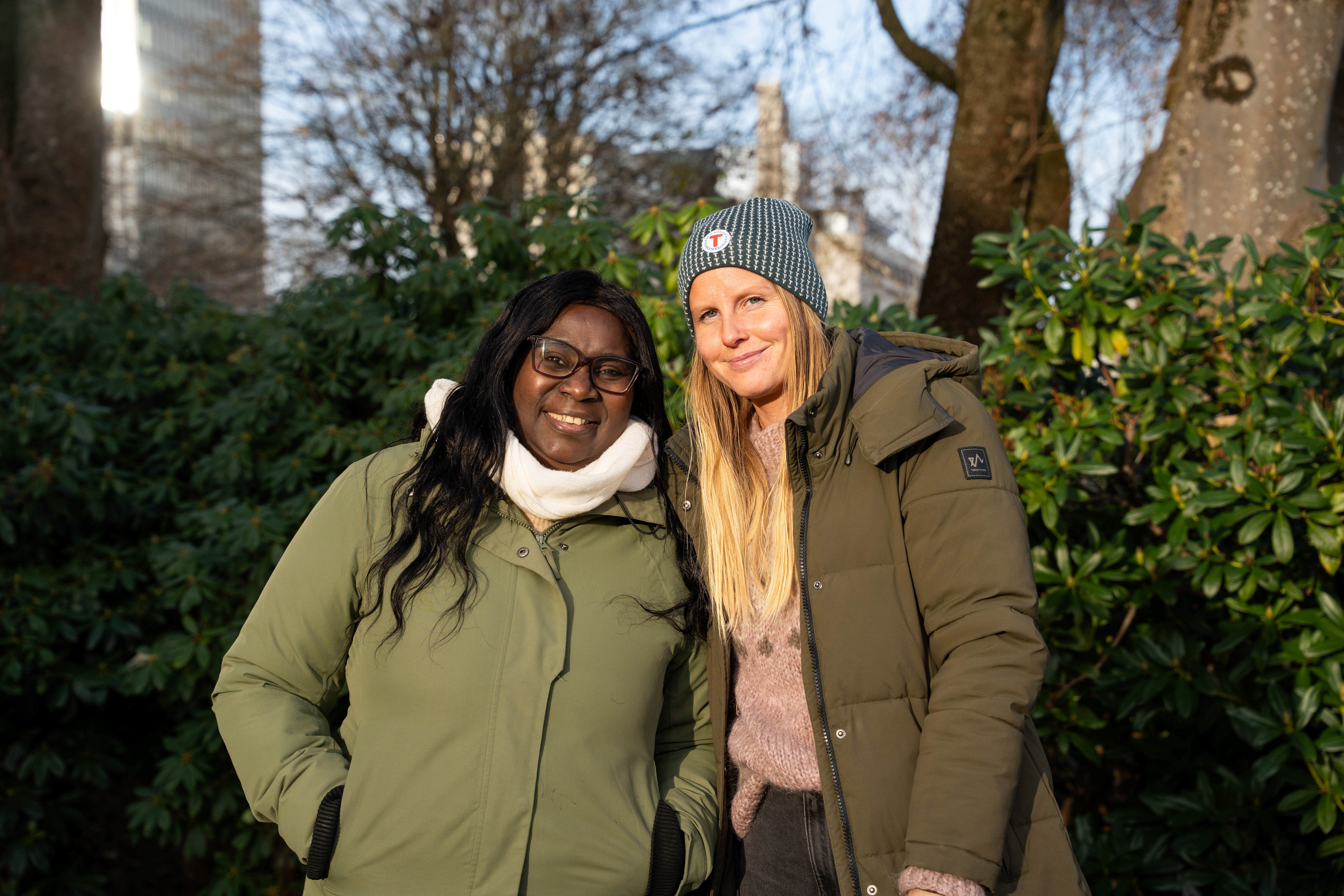 To damer smiler, foran busker og trær i bakgrunnen.