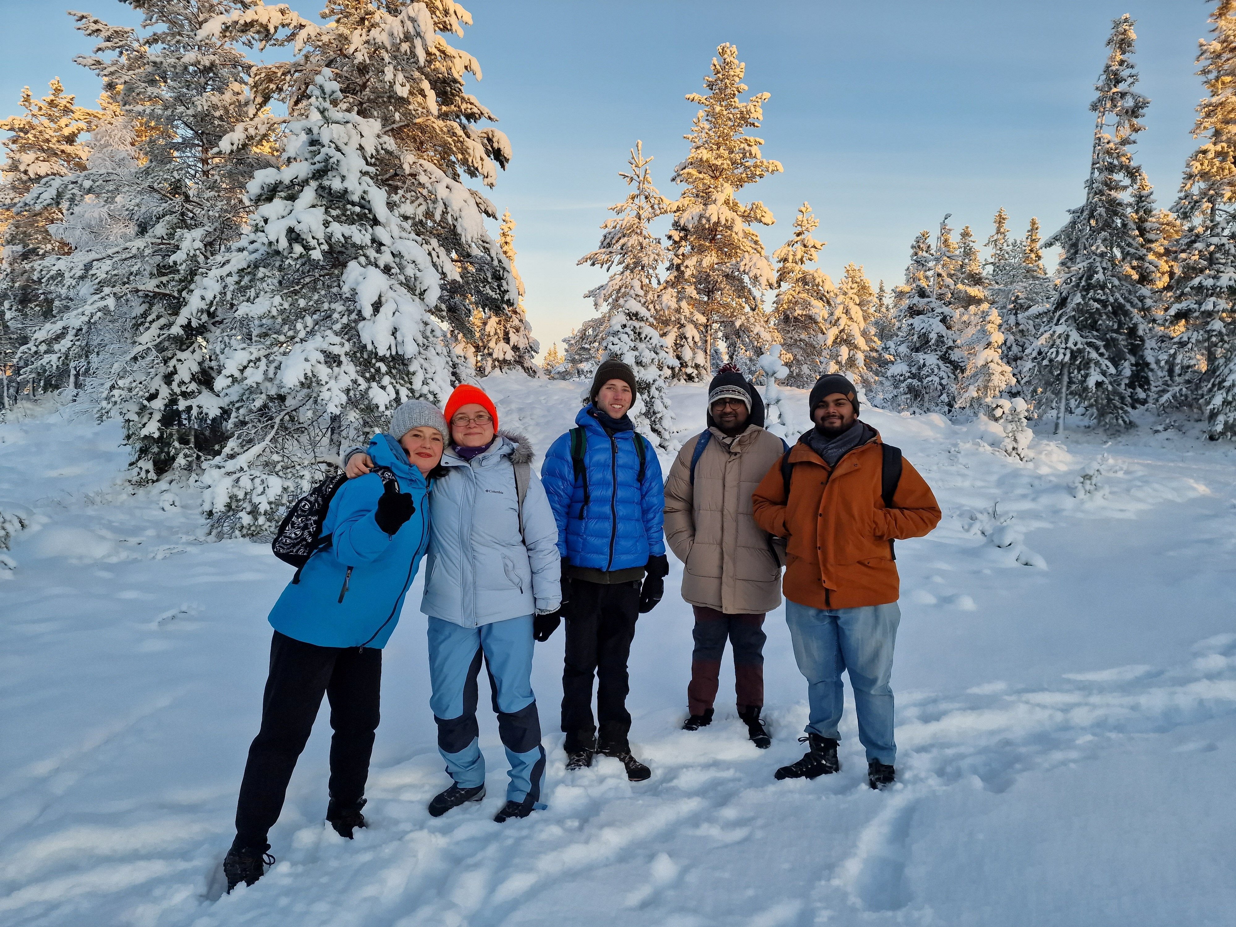 fem personer st&aring;r og ser mot kameraet og smiler. De st&aring;r i et vakkert vinterlandskap med bakken dekket av sn&oslash; og sn&oslash;kledde grantr&aelig;r.