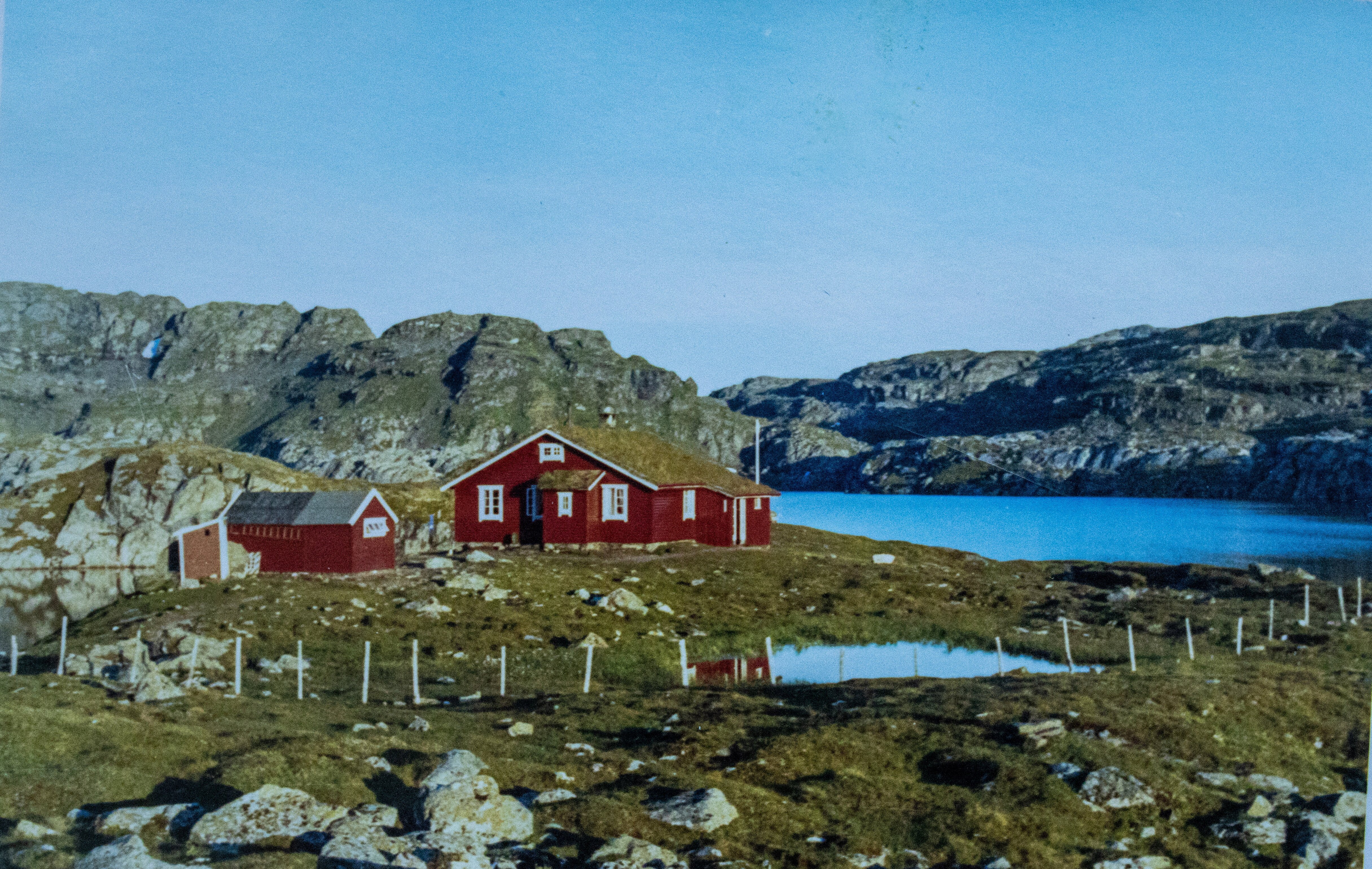 Bilde av en r&oslash;d hytte som ligger ved et vann i storsl&aring;tte naturomgivelser p&aring; fjellet.