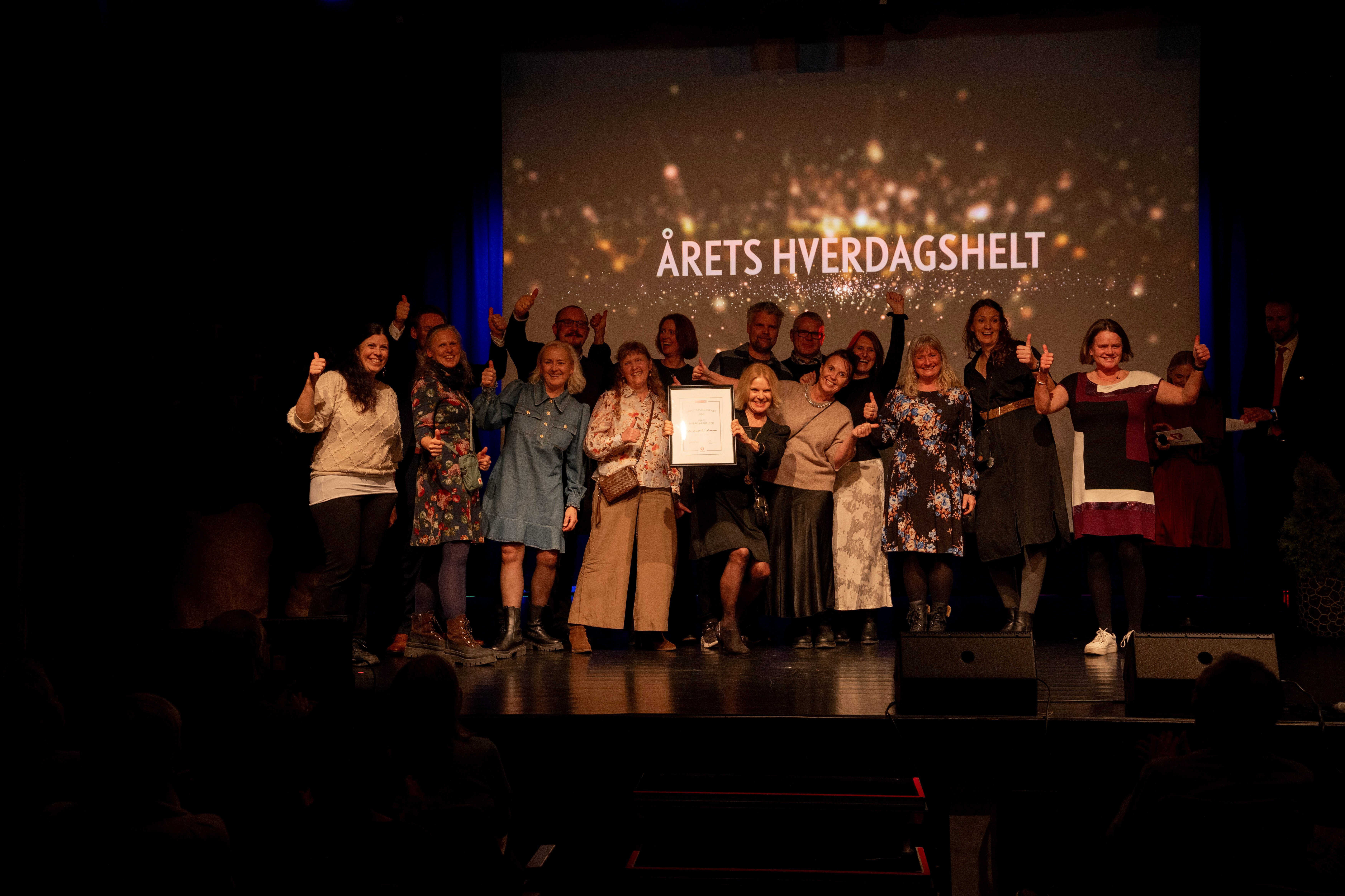 En stor gjeng med festpynta folk står på en scene og smiler til kamera. De har vunnet en pris.