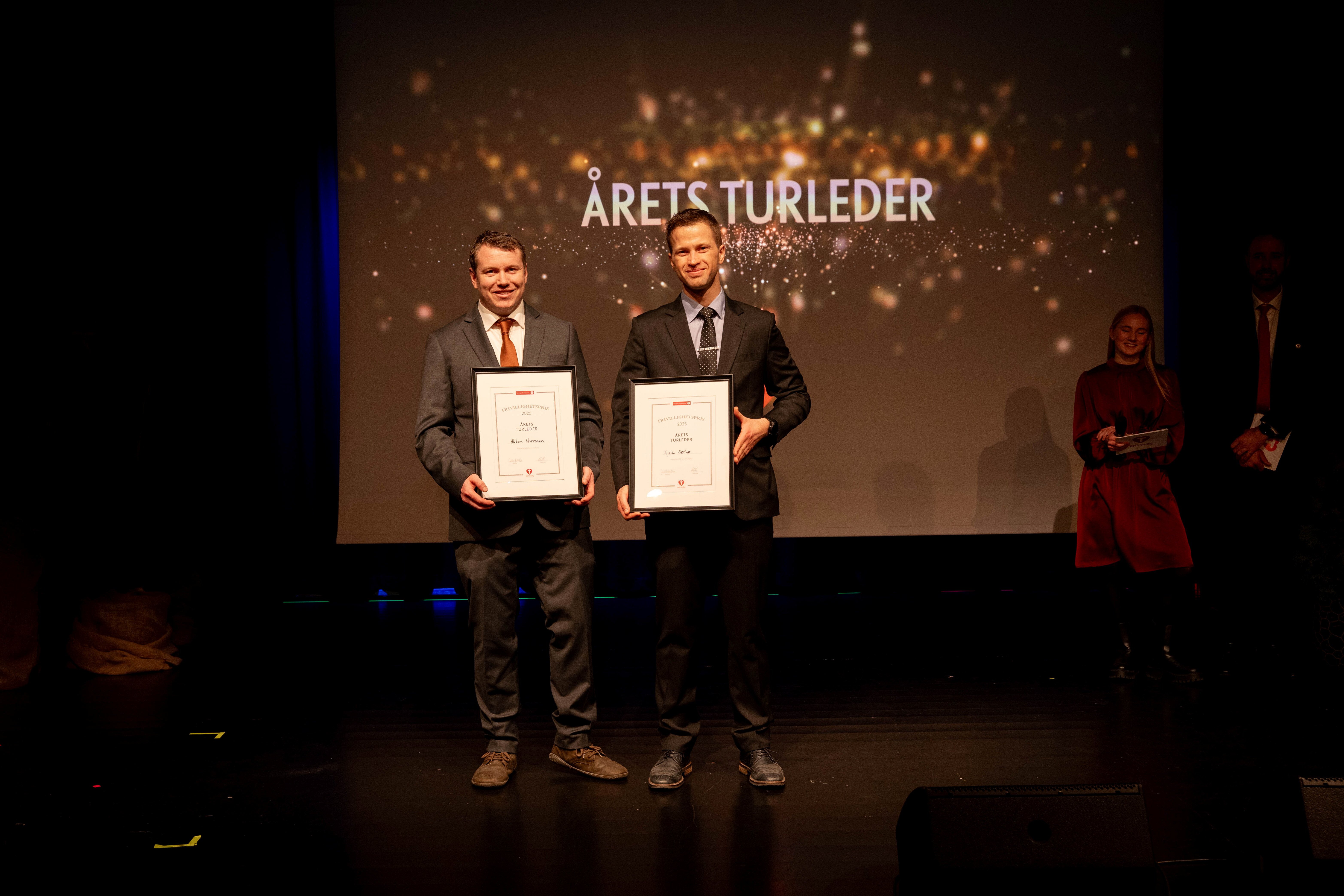 To personer holder fram hver sin ramme med en frivilligpris på en scene.