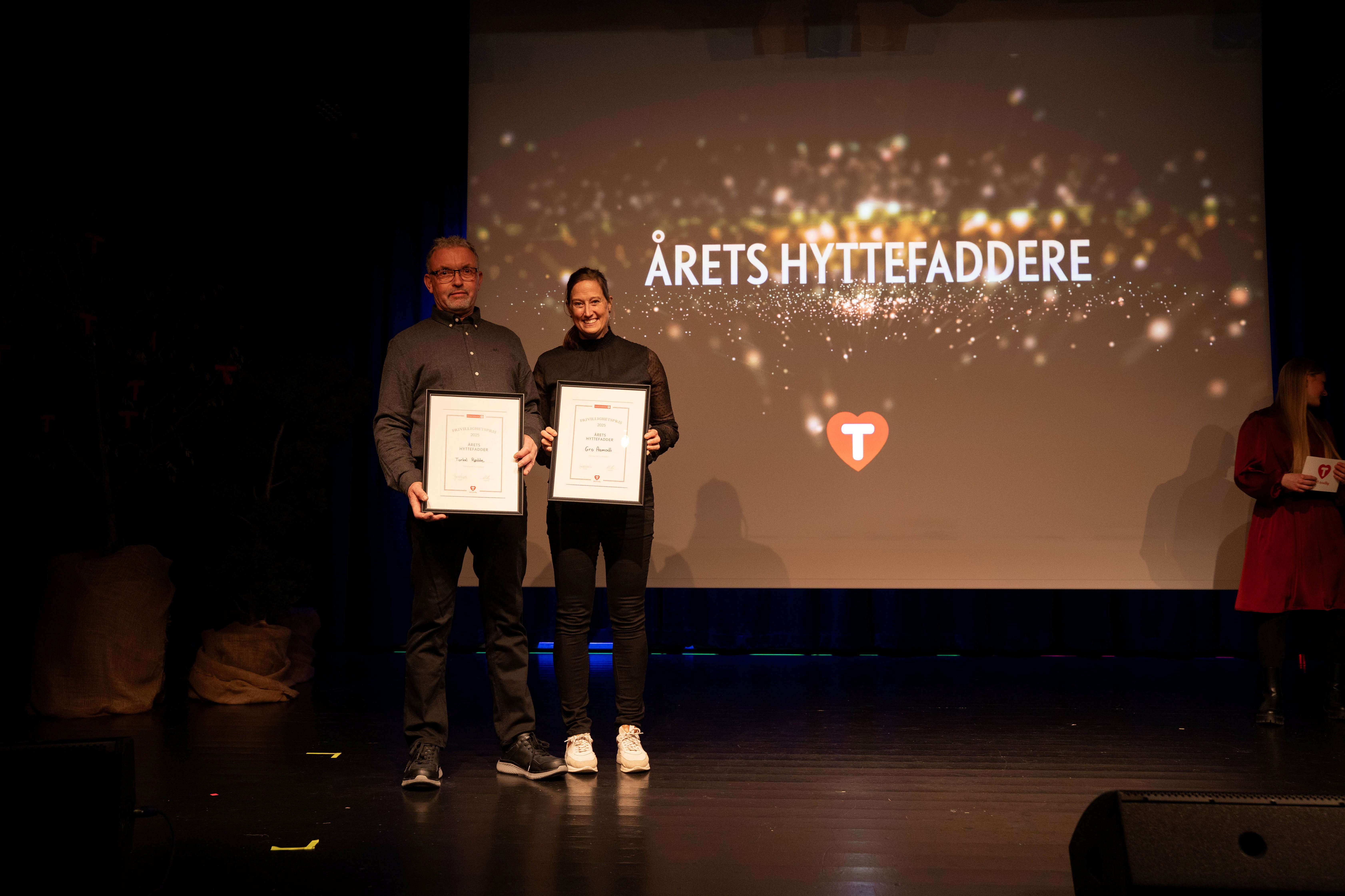 To personer holder fram hver sin ramme med en frivilligpris på en scene.