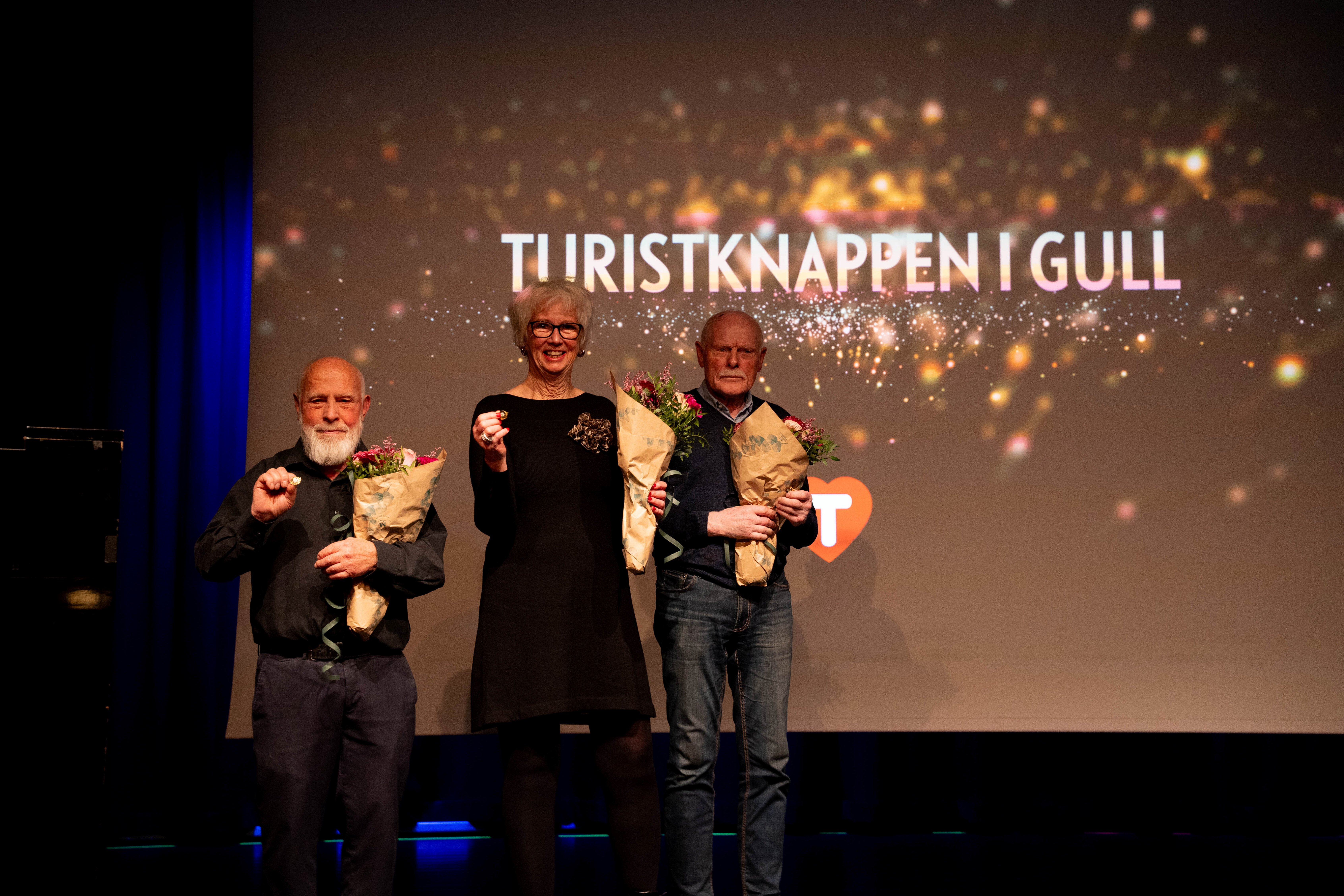 Tre personer står på en scene og holder fram en gullknapp og blomster.