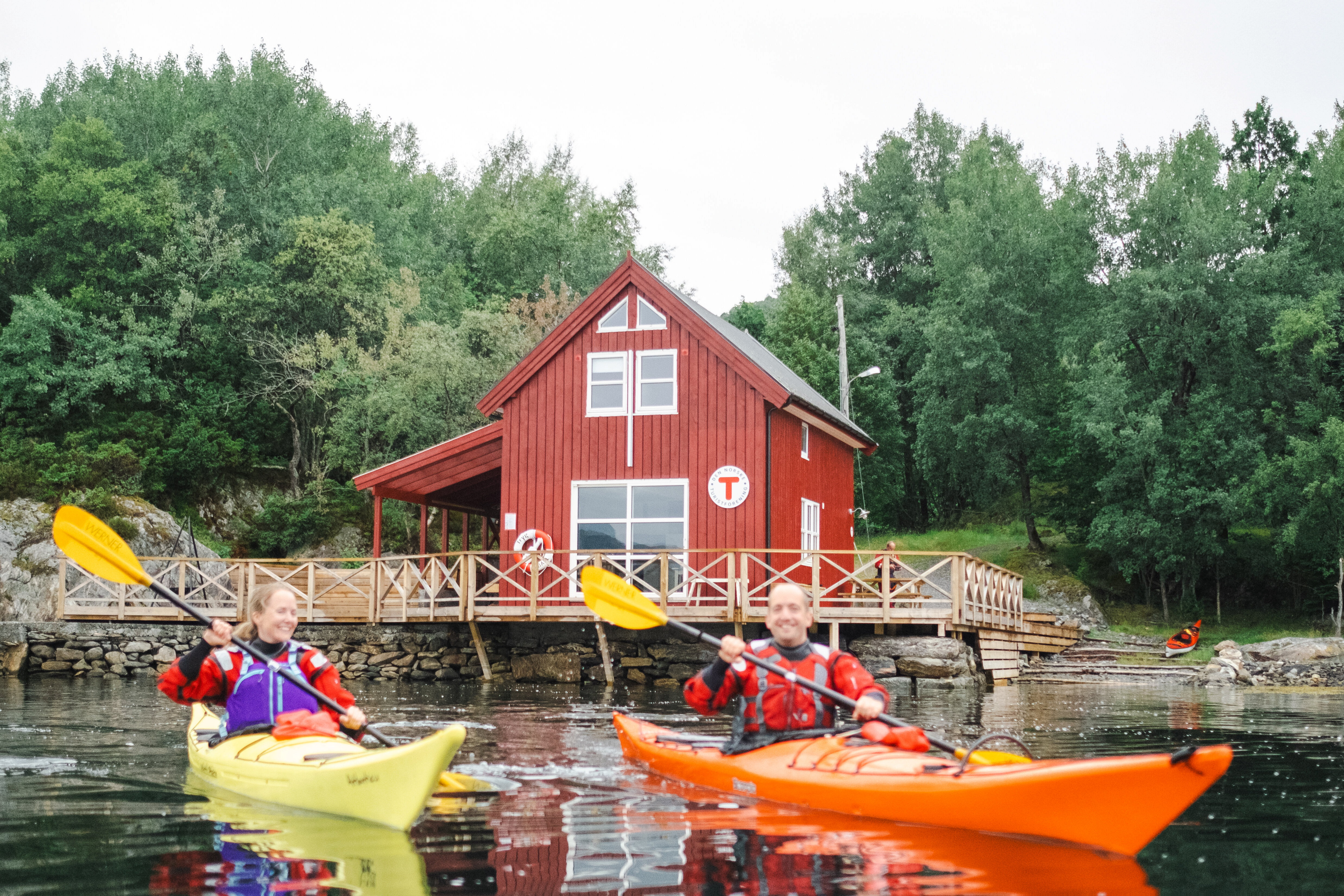 To kajakkpadlere utenfor r&oslash;d hytte som ligner p&aring; en liten sj&oslash;bod.