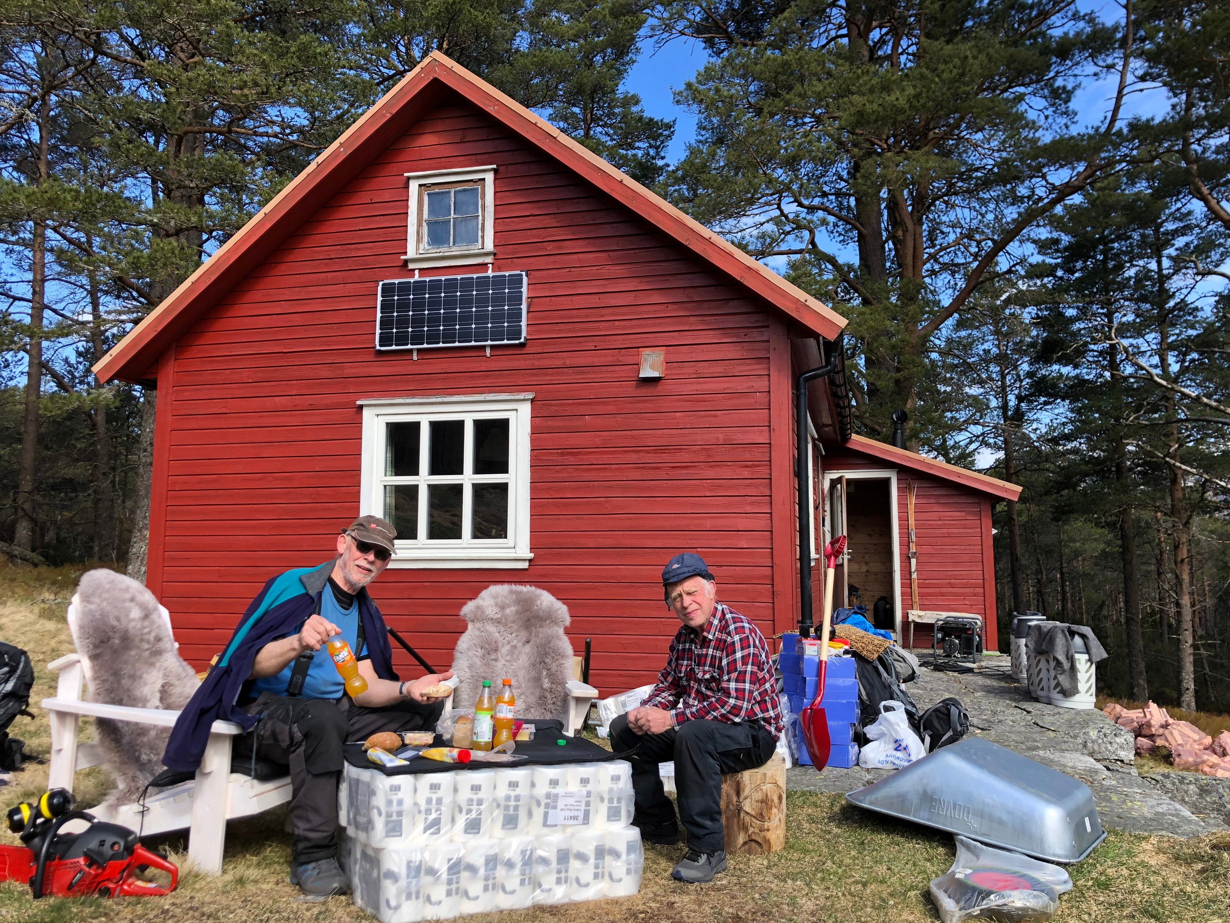 To menn sitter utenfor en rød hytte og spiser lunsj på en dugnad.