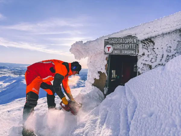 En mann spar bort snø og is utenfor en hytte på fjellet