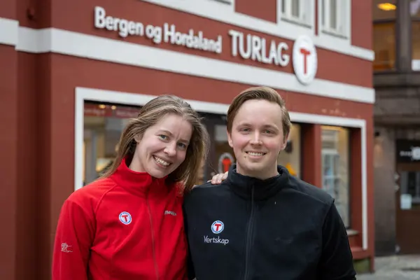 Kvinne i rød genser og mann i blå genser smiler til kamera for Turlagets butikk i Bergen.