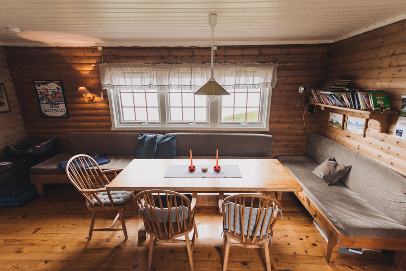 Et hyttestue med vindu på veggen. Et trebord med stoler rundt.  Det står noen grå sofaer langs veggen. 