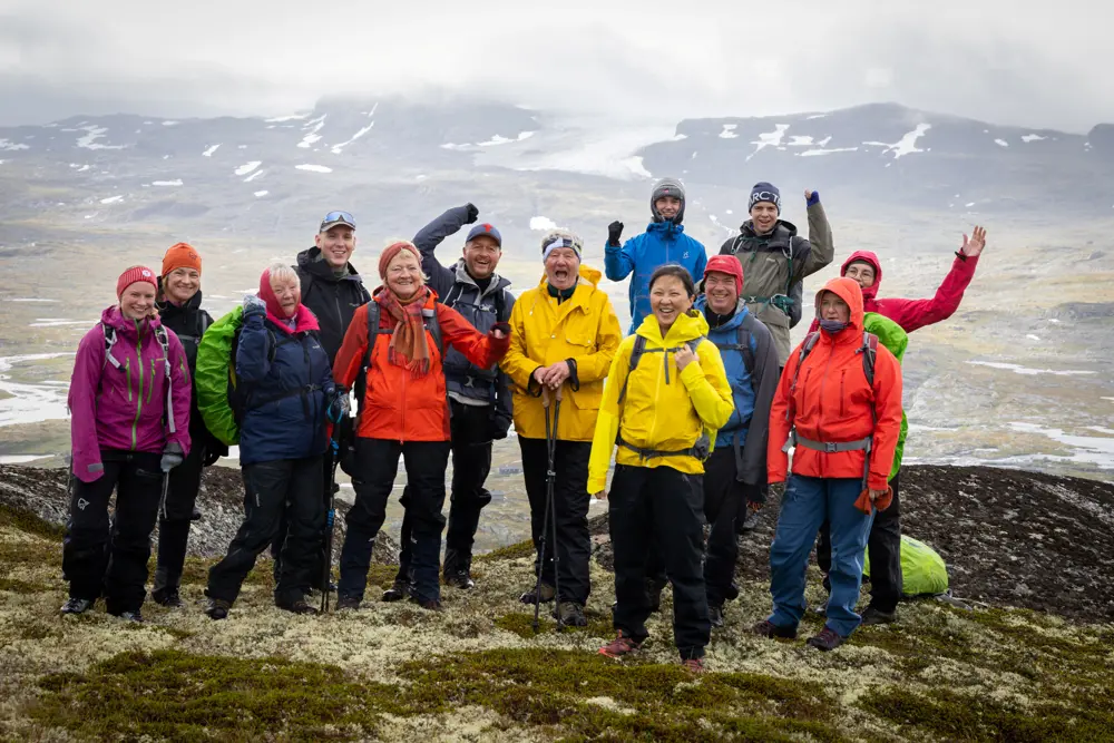 En turgruppe smiler og hoier på fjelltopp med skoddekledde fjell i bakgrunnen på høyfjellet.