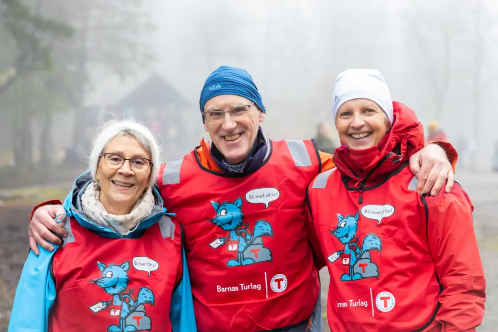 Tre frivillige med røde vester fra Turistforeningen smiler til kamera, en mann i midten som holder rundt to damer.