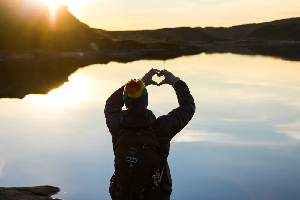 Kvinne i siluett folder hendene til et hjerte foran solnedgang ved et stille vann og fjell bak.