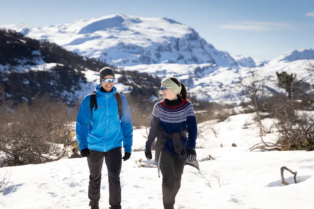 Mann i blå jakke og kvinne i mariusgenser smiler og går på tur i fjellet med snø og lyng.