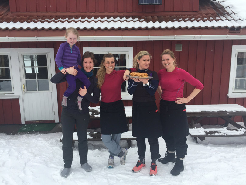 Fire mennesker står foran en hytte. En har et barn på skulderen