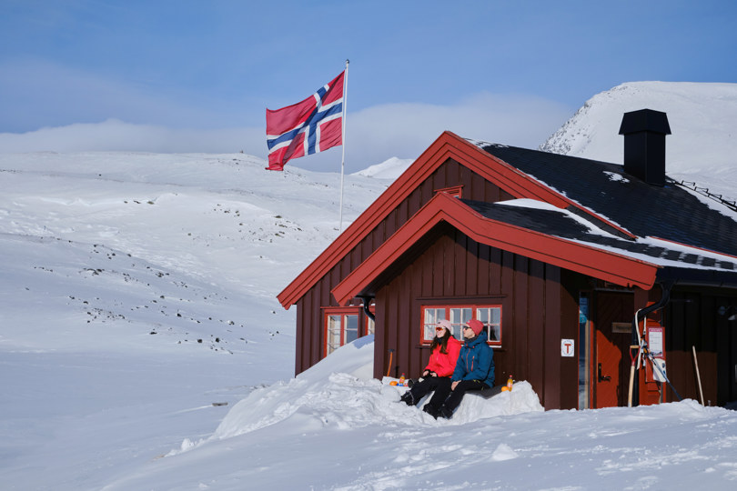 To mennesker i solveggen utenfor DNT-hytte på fjellet. Vinter